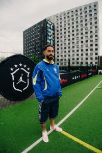 Matheus Cunha standing on the pitch in a blue Jordan Brasil jacket beside the Jordan Brasil crest banner