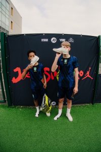 Two young players in blue Brasil kits holding up their new Jordan trainers in front of the Joga Sinistro backdrop