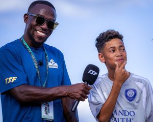 A TSF coach in a blue TSF shirt holding a TSF microphone up to a smiling young player in an Orlando City kit