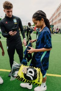 A young player looking through the Brasil kit from her yellow JD Sports bag while a coach films on his phone